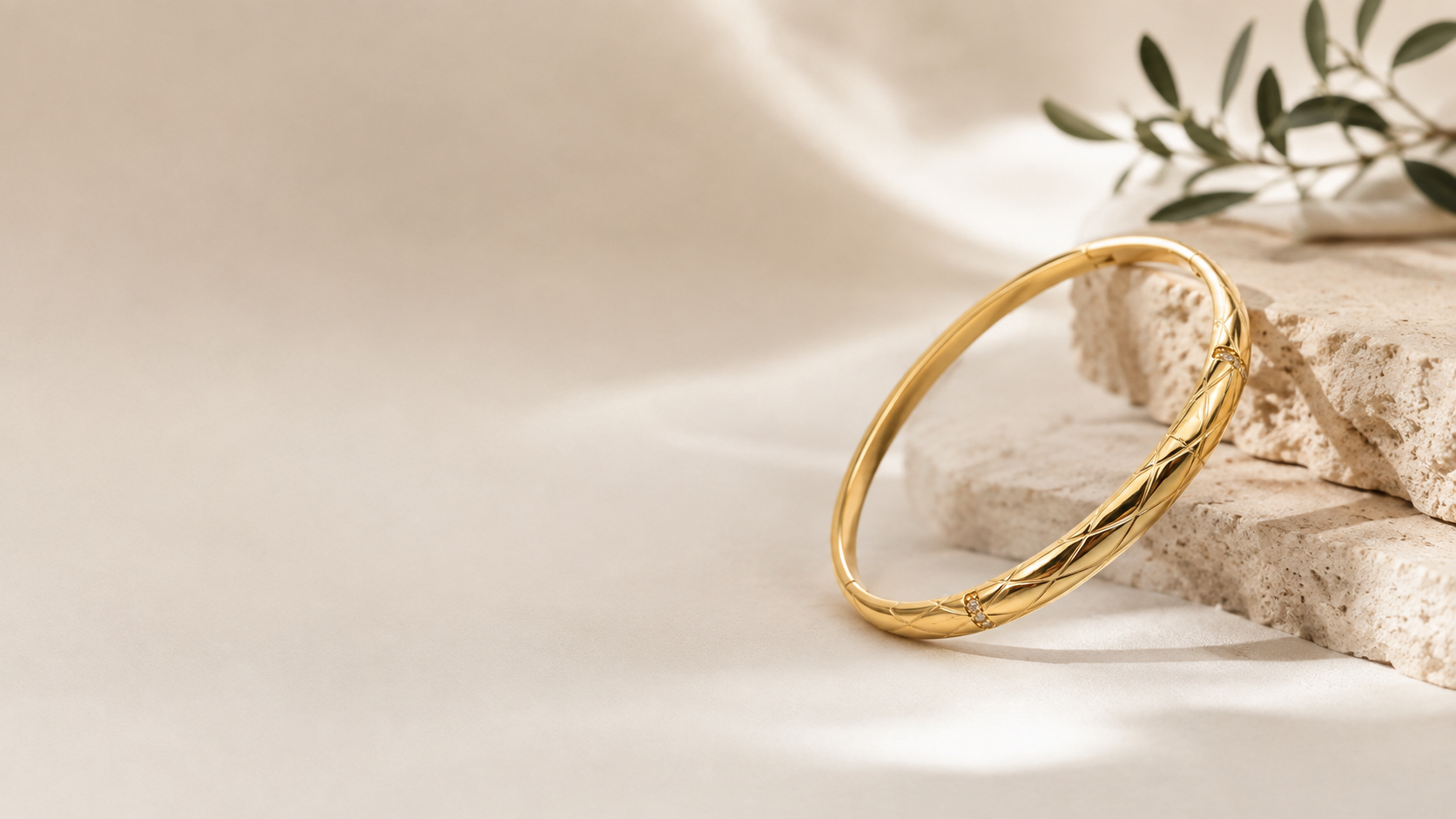 A polished gold bangle bracelet with a twisted, textured design, resting on stacked beige stone blocks with soft shadows and a small green leafy sprig in the background.